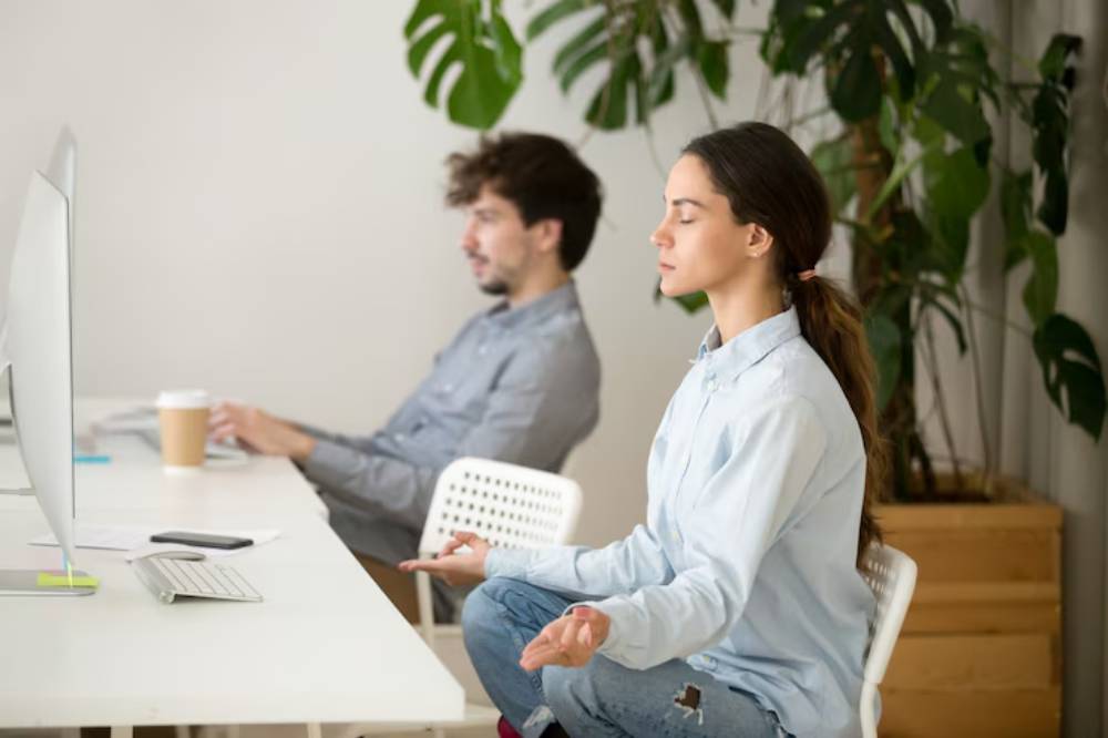 A woman meditates in a modern office, seated cross-legged at a desk.